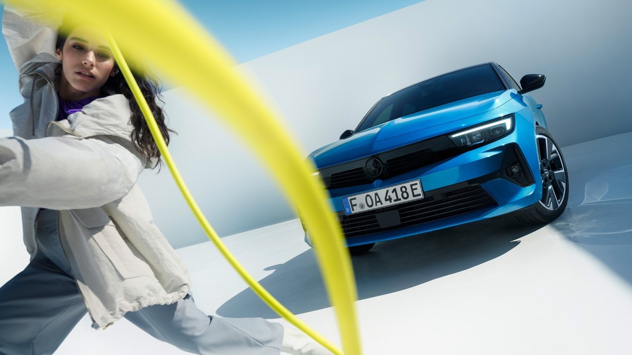 Frontansicht eines blauen Opel Astra Electric mit einer Frau, die ein gelbes Ladekabel herumwirbelt