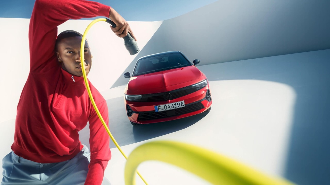 Ein Mann hält ein Ladekabel in der Hand und steht vor einem roten Opel Astra Sports Tourer mit schwarzem Dach